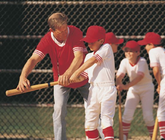 Coach in a red shirt instructing a young baseball player in a white and red uniform on batting technique, with other young players practicing in the background. Genesis Sports Therapy of Hickory, NC, offers LED light therapy to accelerate an athlete's recovery and enhance performance with targeted LED light therapy.