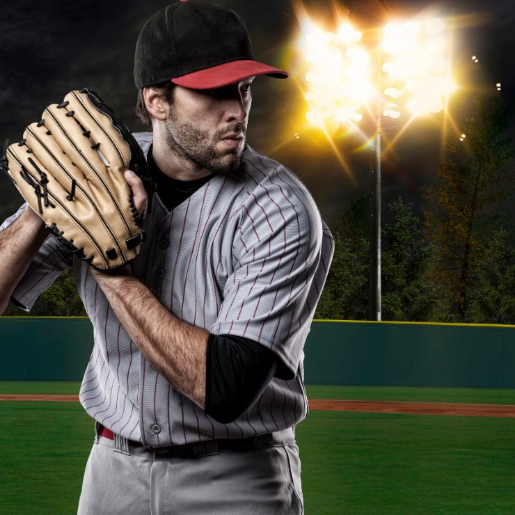 A focused baseball pitcher in a striped uniform and cap, poised to throw a pitch under bright stadium lights. This image highlights how Genesis Sports Therapy, of Hickory, NC offers LED light therapy to enhance athletes' endurance and peak performance during intense games.