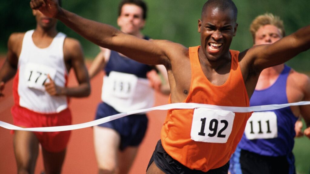 A determined athlete wearing an orange jersey and bib number 192 crosses the finish line first in a track race, arms outstretched in victory, followed closely by other competitors. Genesis Sports Therapy of Hickory, NC, offers LED light therapy to accelerate an athlete's recovery and enhance performance with targeted LED light therapy.