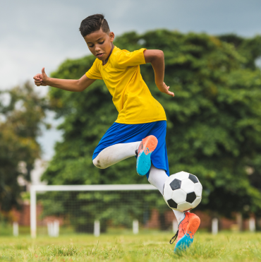 Young soccer player in a yellow shirt and blue shorts mid-air, kicking a soccer ball on a grassy field with trees in the background. Genesis Sports Therapy of Hickory, NC, offers LED light therapy to accelerate an athlete's recovery and enhance performance with targeted LED light therapy.