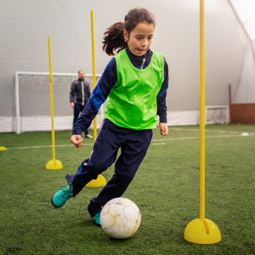 Young girl in a green training vest and blue tracksuit dribbling a soccer ball around yellow cones during a soccer practice in an indoor facility. Genesis Sports Therapy of Hickory, NC, offers LED light therapy to accelerate an athlete's recovery and enhance performance with targeted LED light therapy.