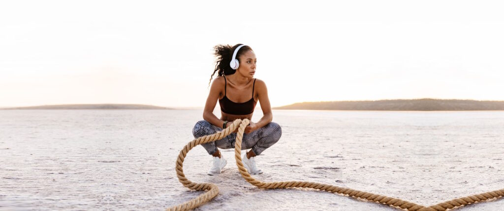 A fit woman in a sports bra and leggings, wearing headphones, squats on a sandy landscape while holding heavy ropes, ready for an intense workout session at dawn. Genesis Sports Therapy of Hickory, NC, offers LED light therapy to accelerate an athlete's recovery and enhance performance with targeted LED light therapy, helping athletes maintain peak physical condition and recover quickly from rigorous training.