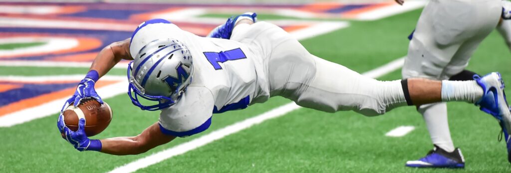 Football player in full gear makes a diving catch mid-air during a game, showcasing athletic performance at its peak. Genesis Sports Therapy, 231 13th Pl NW Suite B Hickory, NC, 28601, specializes in LED light therapy to help athletes like this recover swiftly and perform at their best.