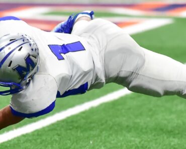Football player in full gear makes a diving catch mid-air during a game, showcasing athletic performance at its peak. Genesis Sports Therapy, 231 13th Pl NW Suite B Hickory, NC, 28601, specializes in LED light therapy to help athletes like this recover swiftly and perform at their best.