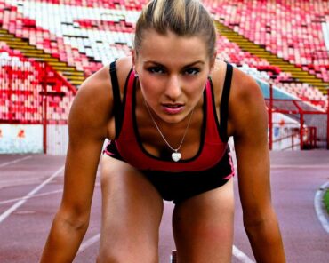 Focused female athlete in starting position on a track, preparing to sprint. She is in an intense, professional setting, emphasizing her dedication and strength. The image was captured at a vibrant stadium with red and white seats in the background, symbolizing competition and passion. This athlete could benefit from Genesis Sports Therapy, 231 13th Pl NW Suite B Hickory, NC, 28601, where LED light therapy enhances peak performance and supports swift recovery for athletes.